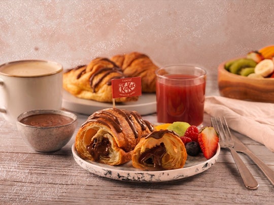 Croissant relleno con chocolate KITKAT® acompañado de frutas, bebida y café en mesa