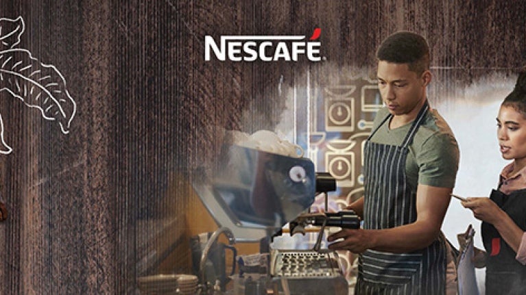 Joven preparando una taza de café en una Máquina Nescafé mientas es observado y evaluado por una mujer joven