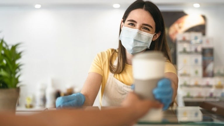 Camarera con delantal, mascarilla de protección y guantes entrega un café a un cliente 