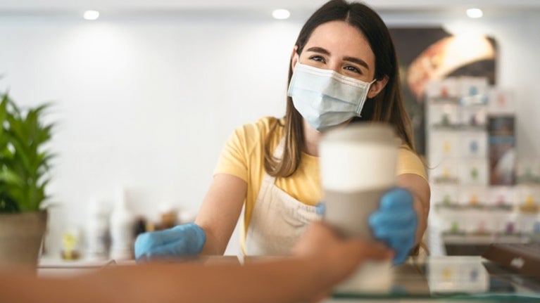 Camarera con delantal, mascarilla de protección y guantes entrega un café a un cliente