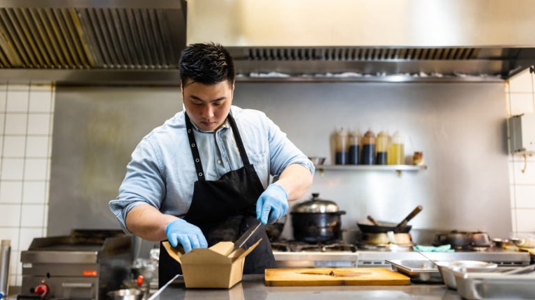En una cocina comercial, chef empaca una preparación en una caja para ser enviada como entrega a domicilio