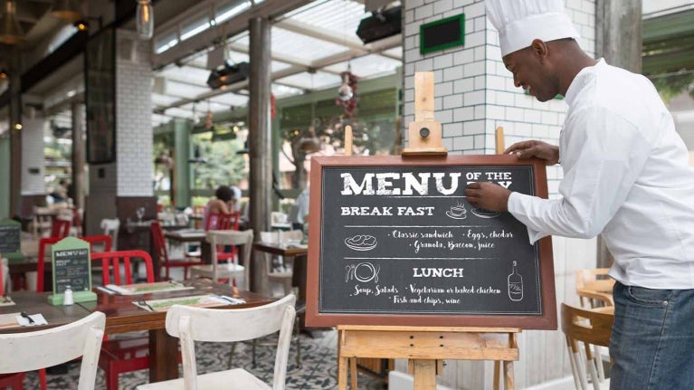 Chef escribe un menú de restaurante sobre una pizarra frente a su negocio que es espacioso y colorido