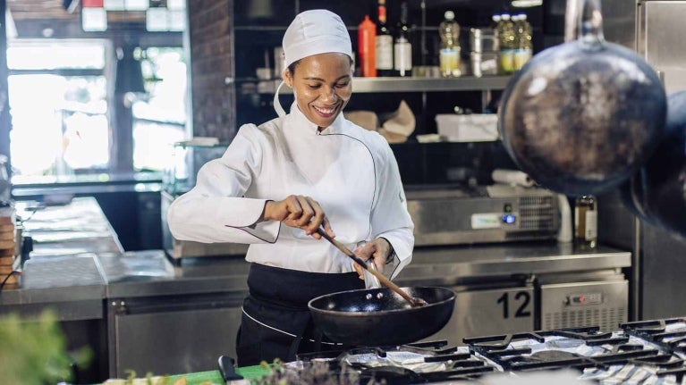 Una chef prepara comida en una sartén, rodeada de un equipamiento de cocina ajustado a las necesidades de un restaurante