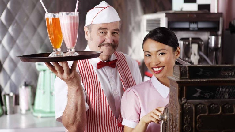 En restaurante temático de los 50, un mesero sostiene una bandeja con jugos y otra opera la caja registradora