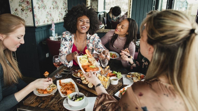 Grupo de 5 amigas sonrientes conmemoran el Día Internacional de la Mujer al compartir un almuerzo especial en un restaurante