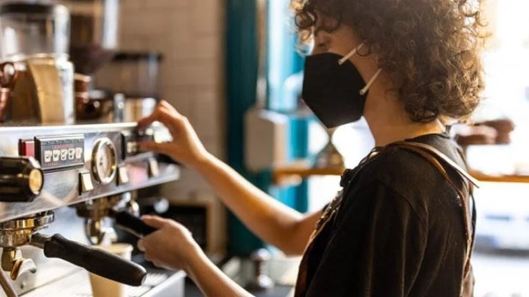 Barista mujer con mascarilla prepara una taza de café en una cafetería