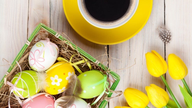 Sobre una mesa de madera 5 rosas amarillas, una taza de café y una canasta con dulces de pascua como huevos decorados