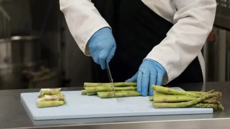 Chef con guantes azules cortando espárragos sobre tabla blanca