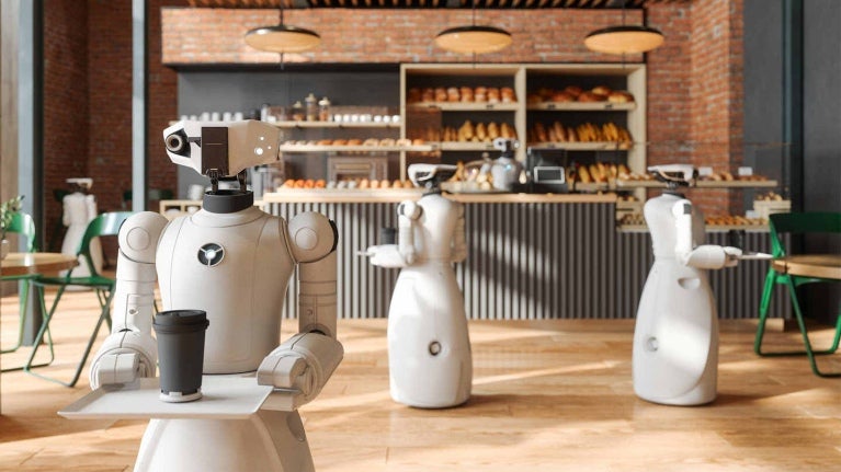 Tres robots recorren una cafetería sosteniendo con sus pinzas una bandeja sobre la que una bebida caliente con tapa