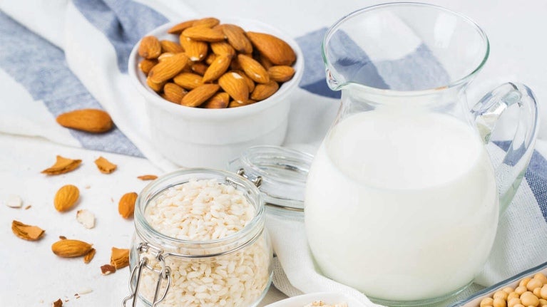 Sobre una mesa una jarra con una bebida vegetal y a su alrededor recipientes con avena, arroz, almendras y garbanzos