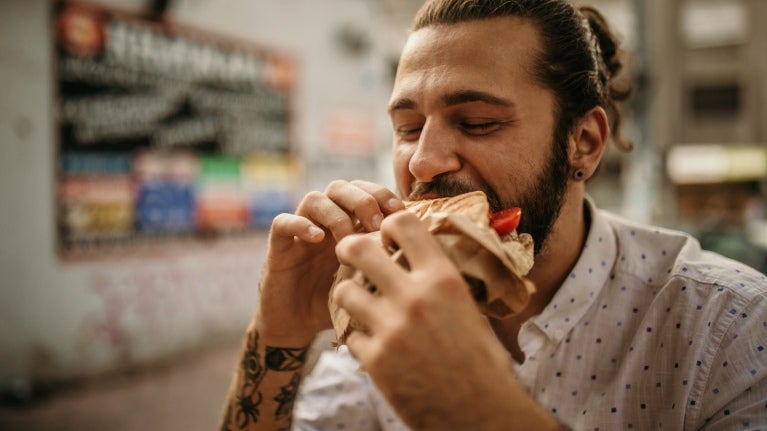 Vista frontal de un hombre que come una arepa rellena, la cual sostiene con sus dos manos, fuera de un negocio de street food