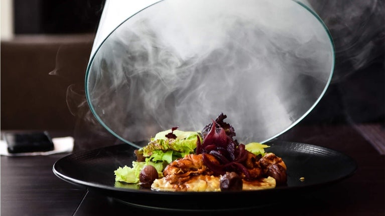 Sobre una mesa de madera se destapa un plato de comida gourmet envuelta en humo