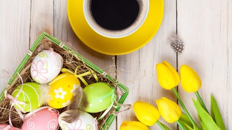 Sobre una mesa de madera 5 rosas amarillas, una taza de café y una canasta con dulces de pascua como huevos decorados