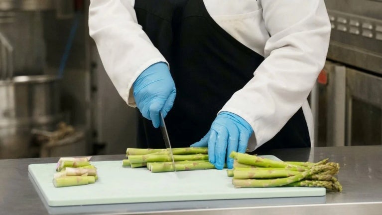 Chef con guantes azules cortando espárragos sobre tabla blanca