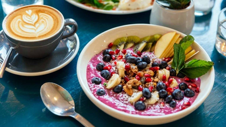 Servido a la mesa un bowl con yogurt y fruta, cappuccino y vasos de agua, que componen un desayuno