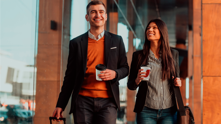 Hombre y mujer sonrientes, caminando en aeropuerto con bebidas de NESCAFÉ en mano y equipaje