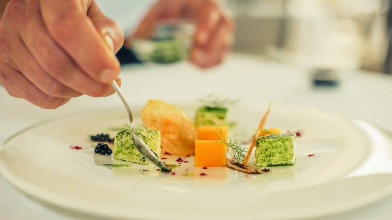 Mano de chef decora con aderezo un plato blanco de vegetales en cubos