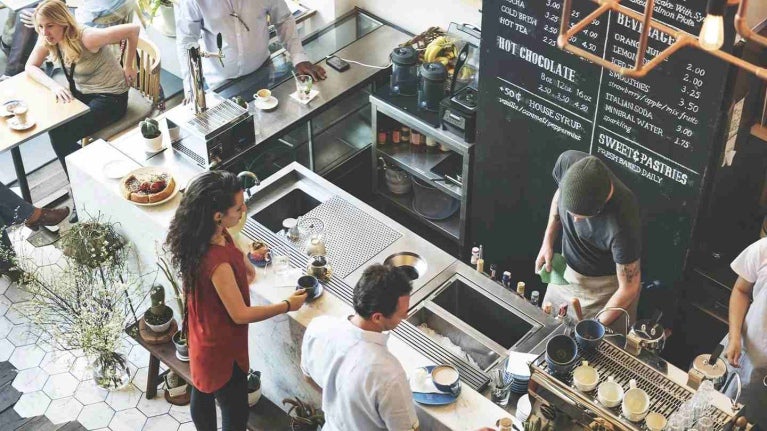 Cafetería concurrida con baristas preparando bebidas y clientes esperando sus pedidos o conversando