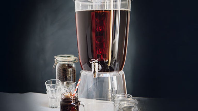 Dispensador de café cold brew junto a vasos y frascos con granos de café en una mesa.