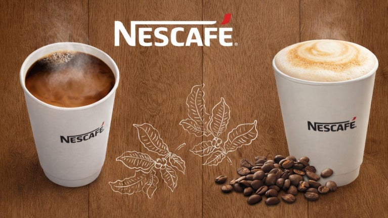 Dos vasos de NESCAFÉ con café americano y cappuccino de calidad, al pie, granos tostados