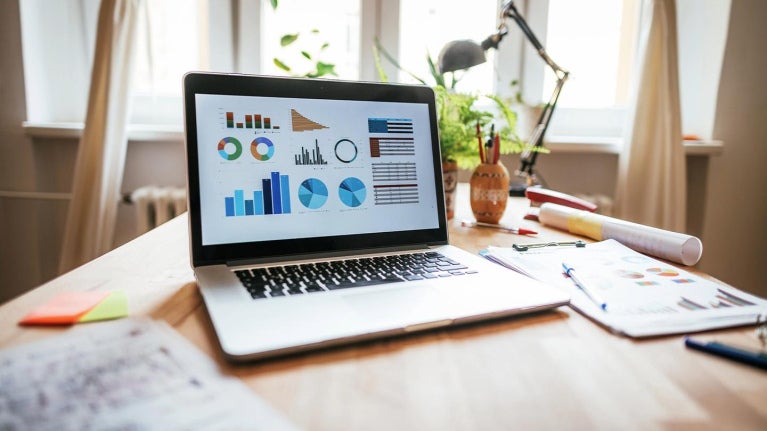 Escritorio con portátil mostrando gráficos y datos, rodeado de documentos, útiles y plantas