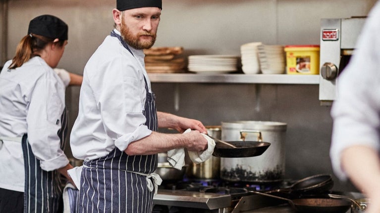 Chef cocinando con sartén frente a estufa en cocina profesional con otros cocineros
