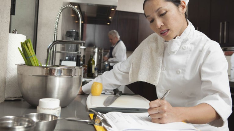 Chef escribiendo en libreta en cocina con utensilios y alimentos sobre mesa