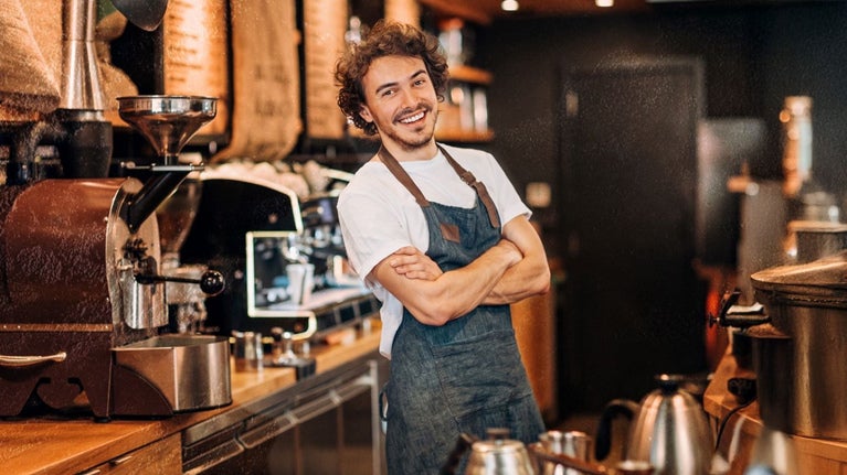Barista sonríe con los brazos cruzados en el área de preparación de una cafetería rodeado de máquinas
