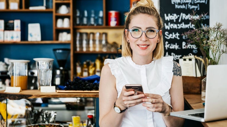 Mujer en cafetería usando celular y laptop, gestionando tareas en entorno de negocio.