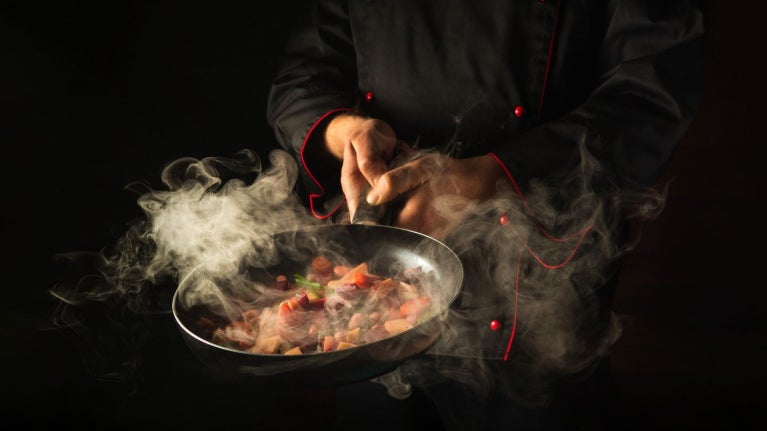 Chef con chaqueta negra sostiene una sartén emanando humo sobre una preparación picada.