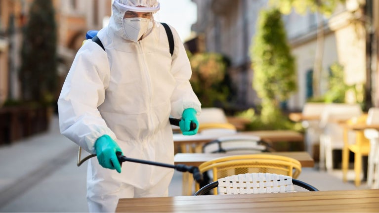Técnico con traje de protección fumiga en la zona de las mesas de un restaurante al aire libre.
