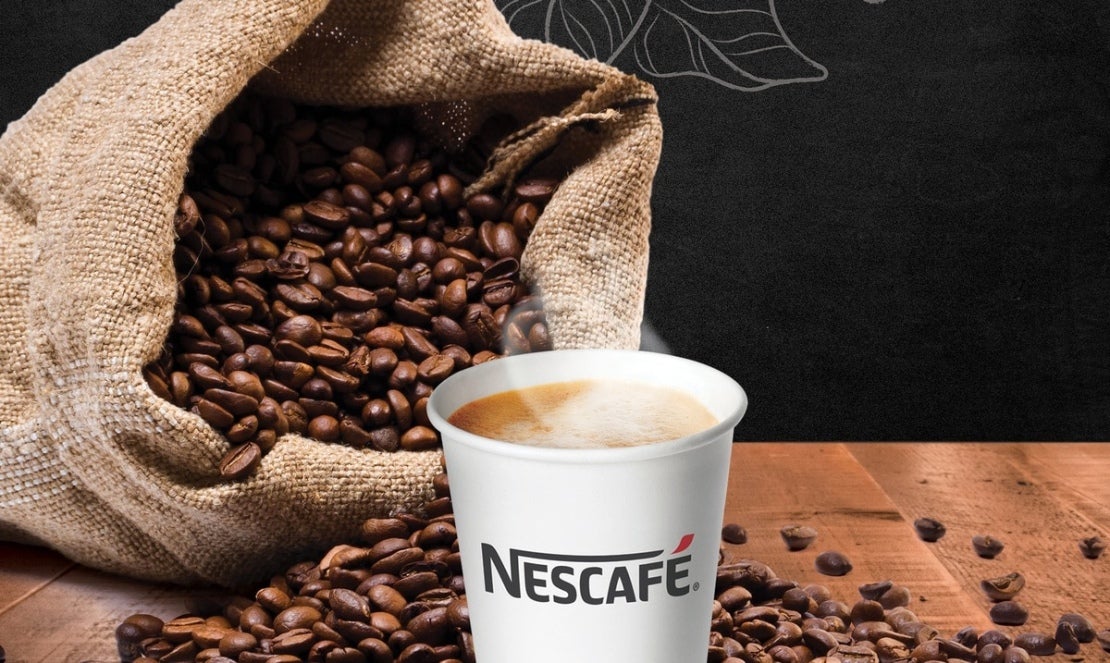 Un vaso de NESCAFÉ® humeante con un costal de granos de café al fondo.