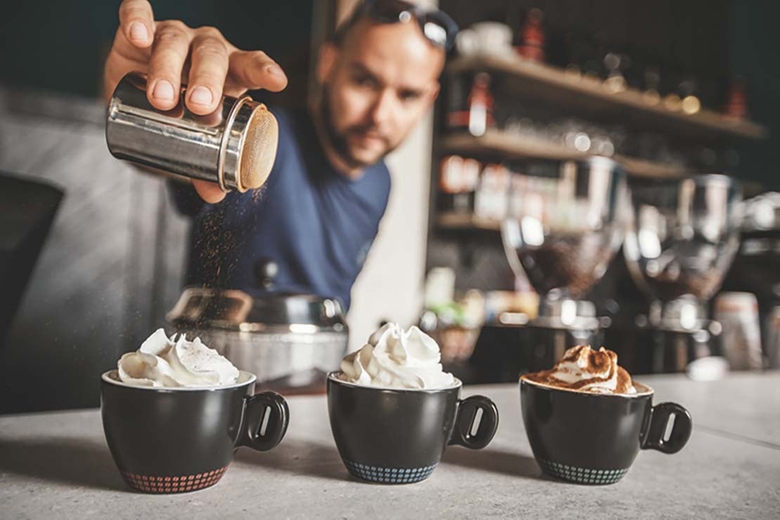 Barista espolvorea cacao sobre tazas de café con nata montada en el Día Mundial del Cacao