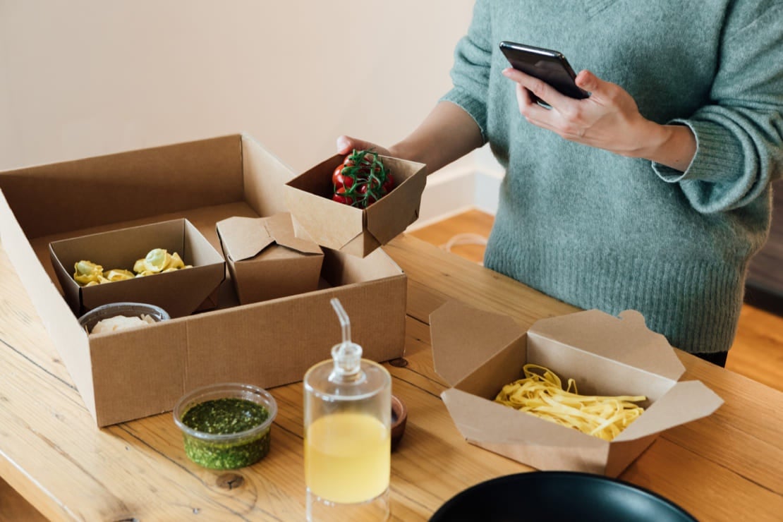 Mujer sostiene un recipiente de comida sacado de una caja de delivery mientras verifica en su celular la entrega a domicilio
