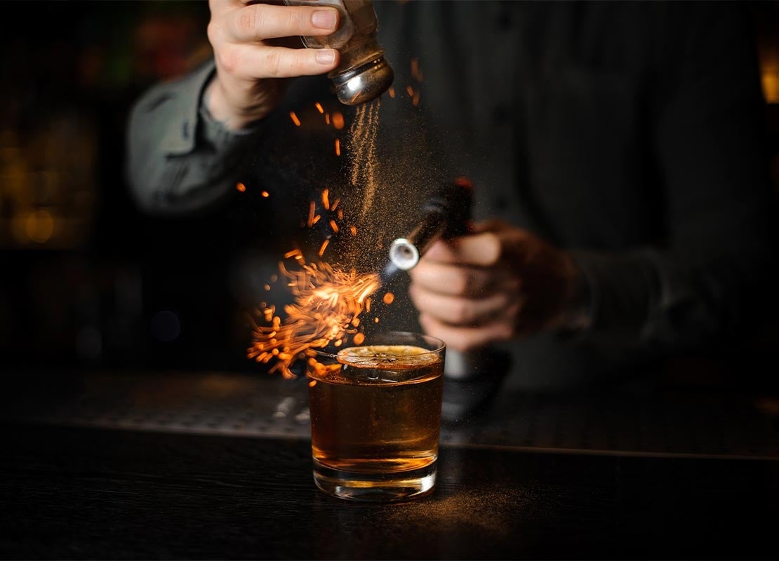Barista añade canela y la flamea con un quemador a un cóctel sin alcohol con una rodaja de naranja seca encima