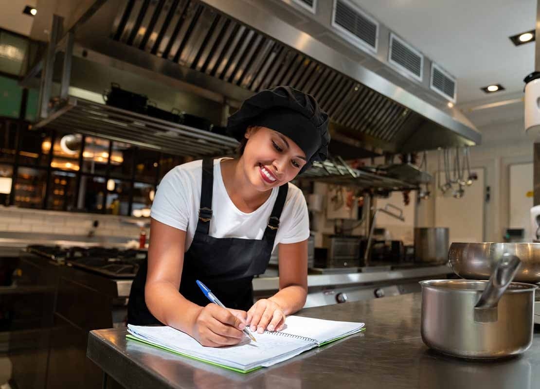 En una cocina comercial, una chef escribe en un cuaderno las descripciones de los platos para el menú de restaurante