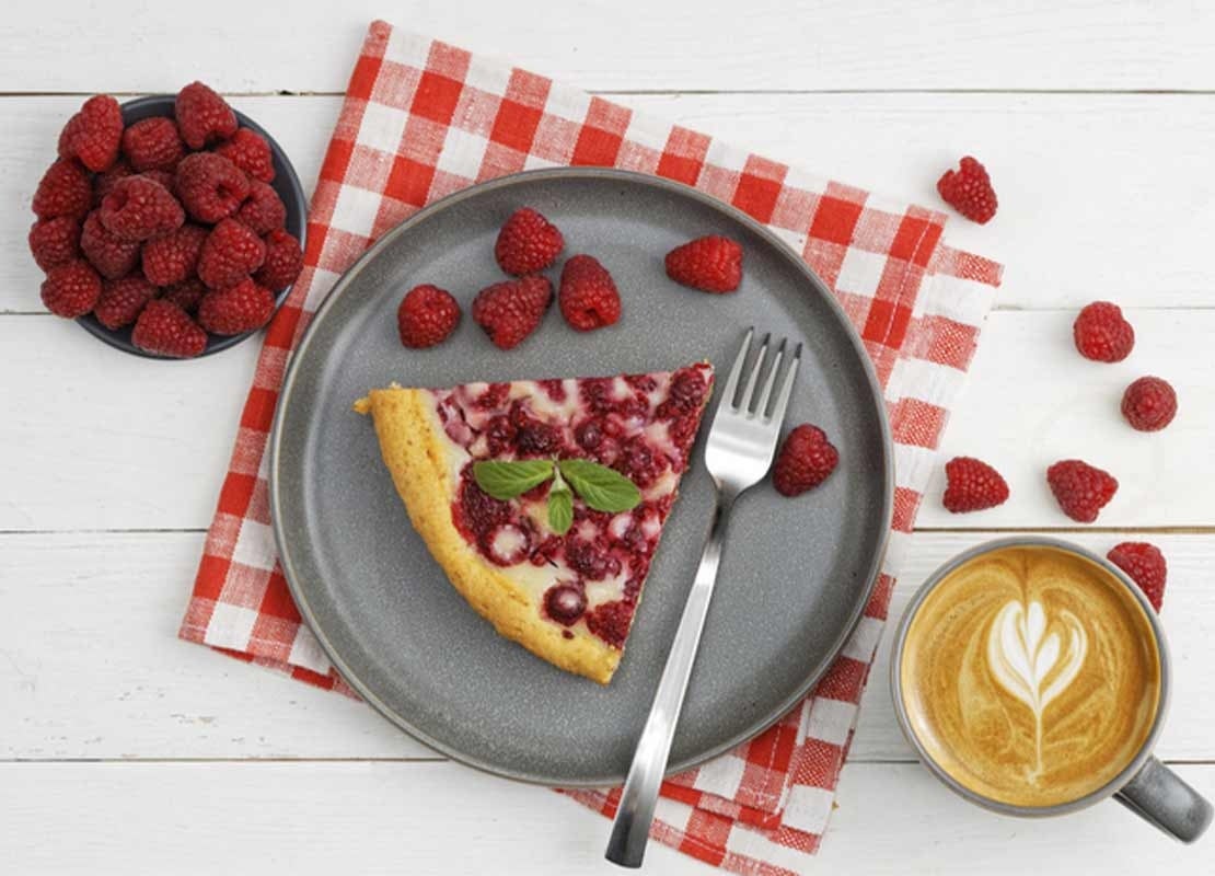 Trozo de tarta de frambuesa, una taza de café y otra de frambuesas dispuestas sobre mesa blanca para food styling