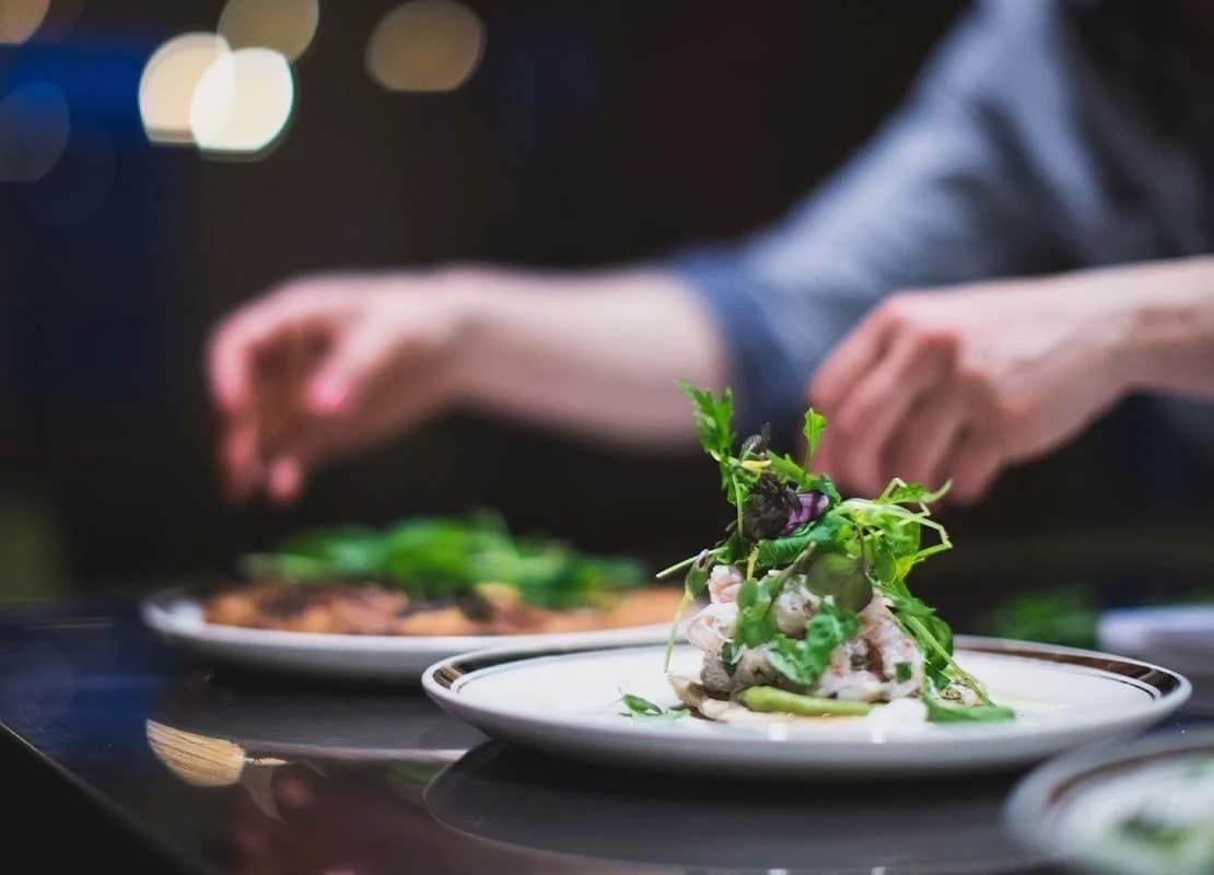 Dos platillos sobre una mesa están siendo decorados y finalizados por un chef en un servicio de catering