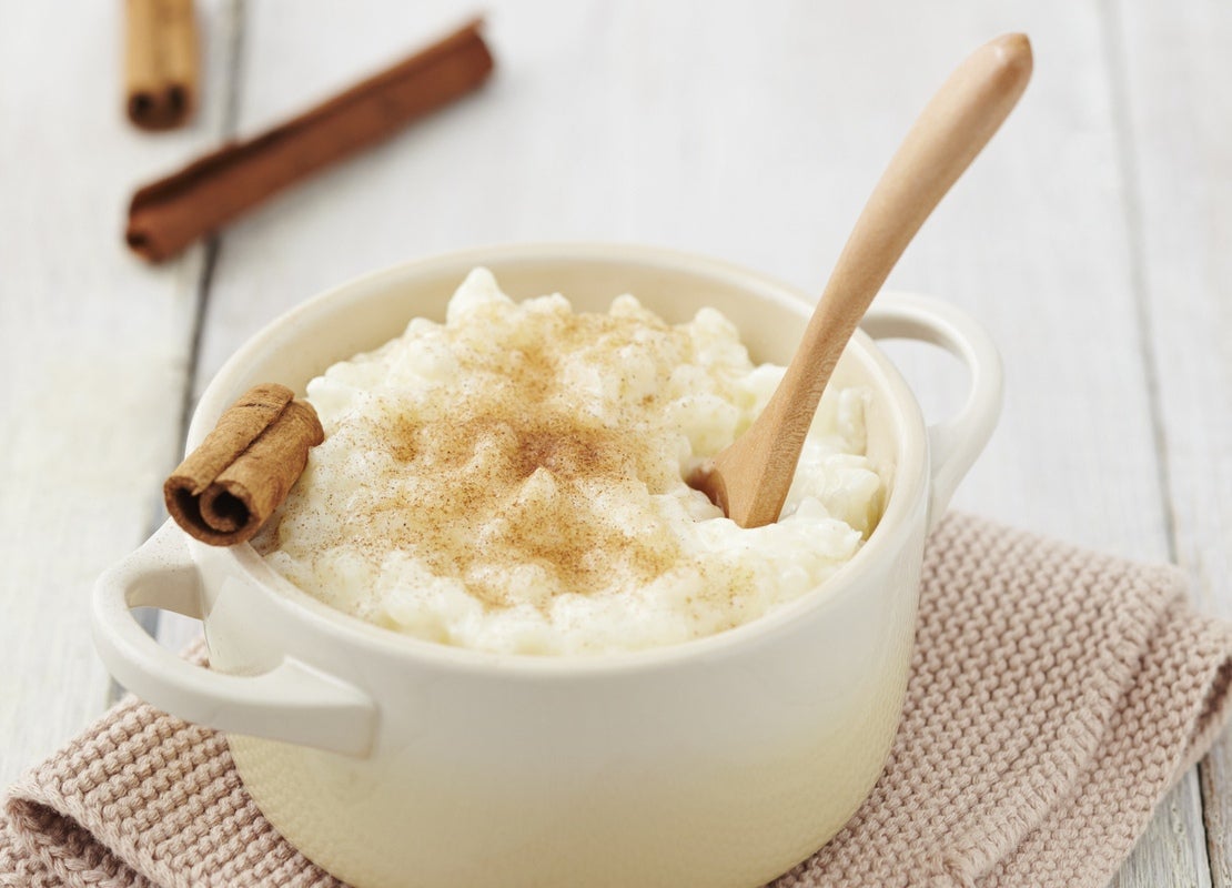 Tazón blanco de arroz con leche decorado con canela en polvo y astilla y una cuchara de madera y servido sobre una tela rosa