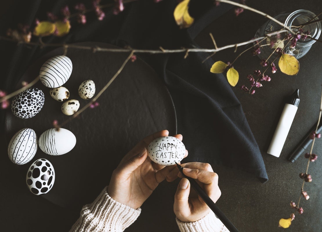 Manos pintan un huevo de pascua con marcador negro y, alrededor, otros huevos decorados y elementos para adornar