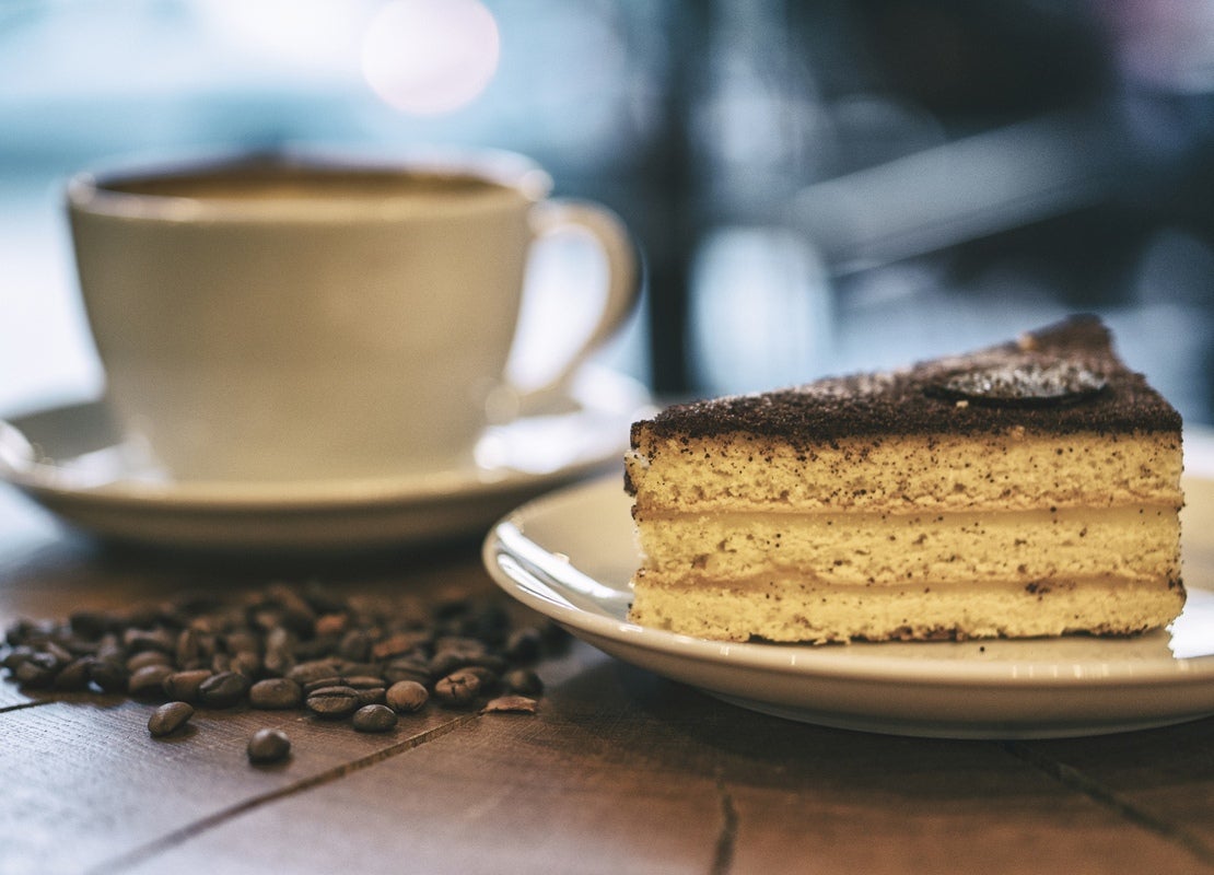 Una taza de espresso y, al frente, un trozo de tarta en una mesa de madera sobre la que hay regados granos de café tostados