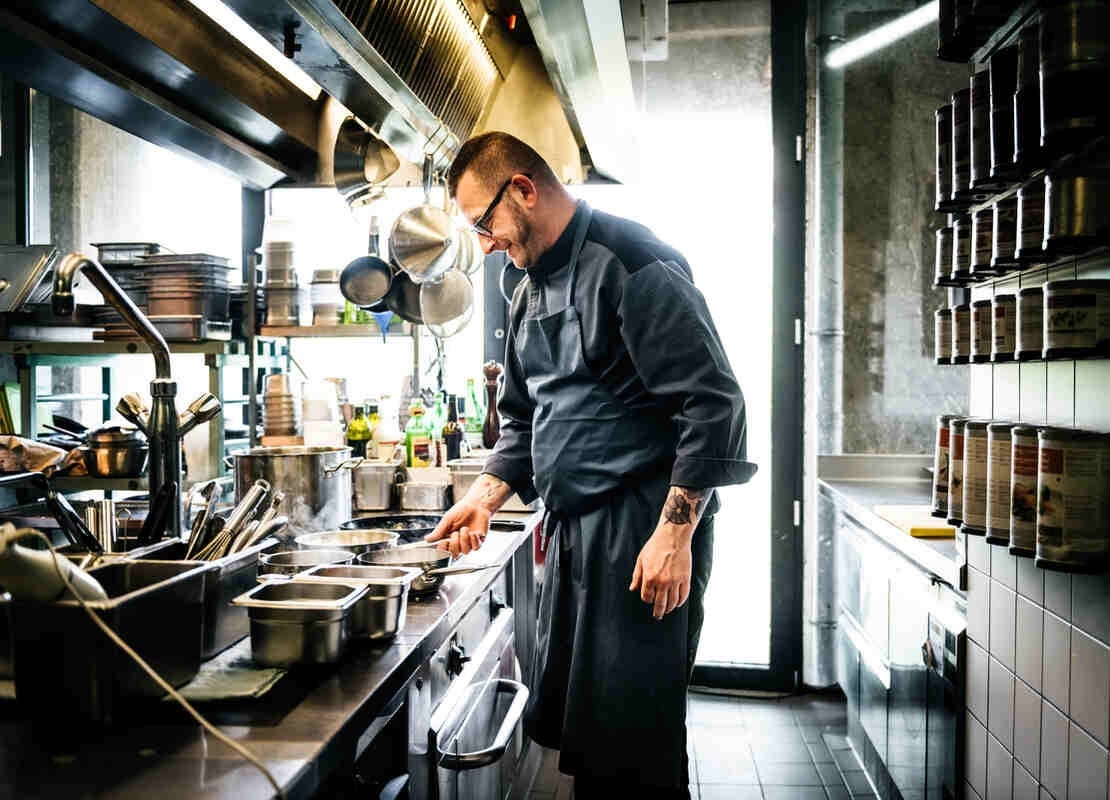Chef revisa ollas que están sobre la estufa de una cocina de restaurante y frente a él hay utensilios en repisas y ganchos