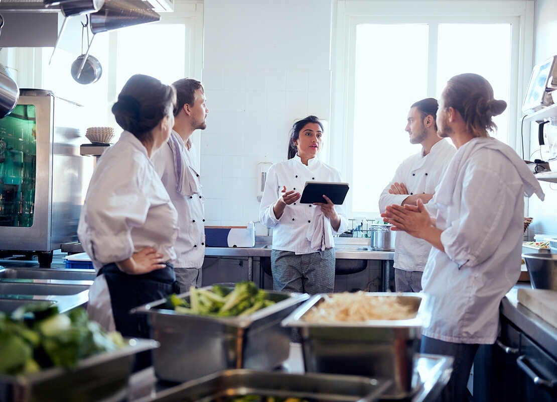 La chef de estación sostiene una Tablet en la mano mientras habla con 4 miembros de su equipo en la cocina del restaurante
