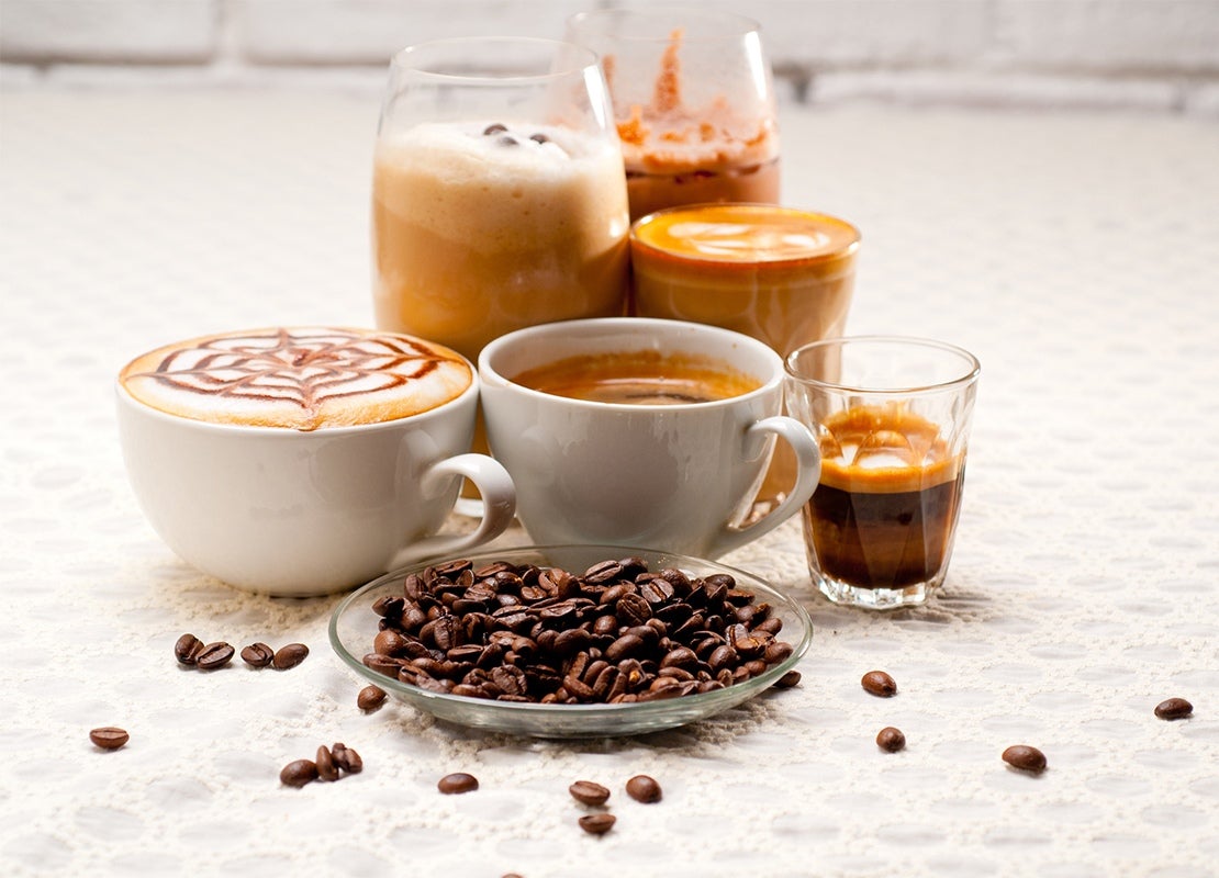 Un plato con granos de café tostados y al fondo seis tipos de bebidas como espresso, café frío, latte, entre otras