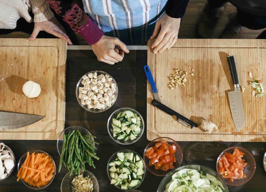 Plano cenital de una mesa con dos tablas de picar sobre las que hay cuchillos y al lado tazones con ingredientes cortados