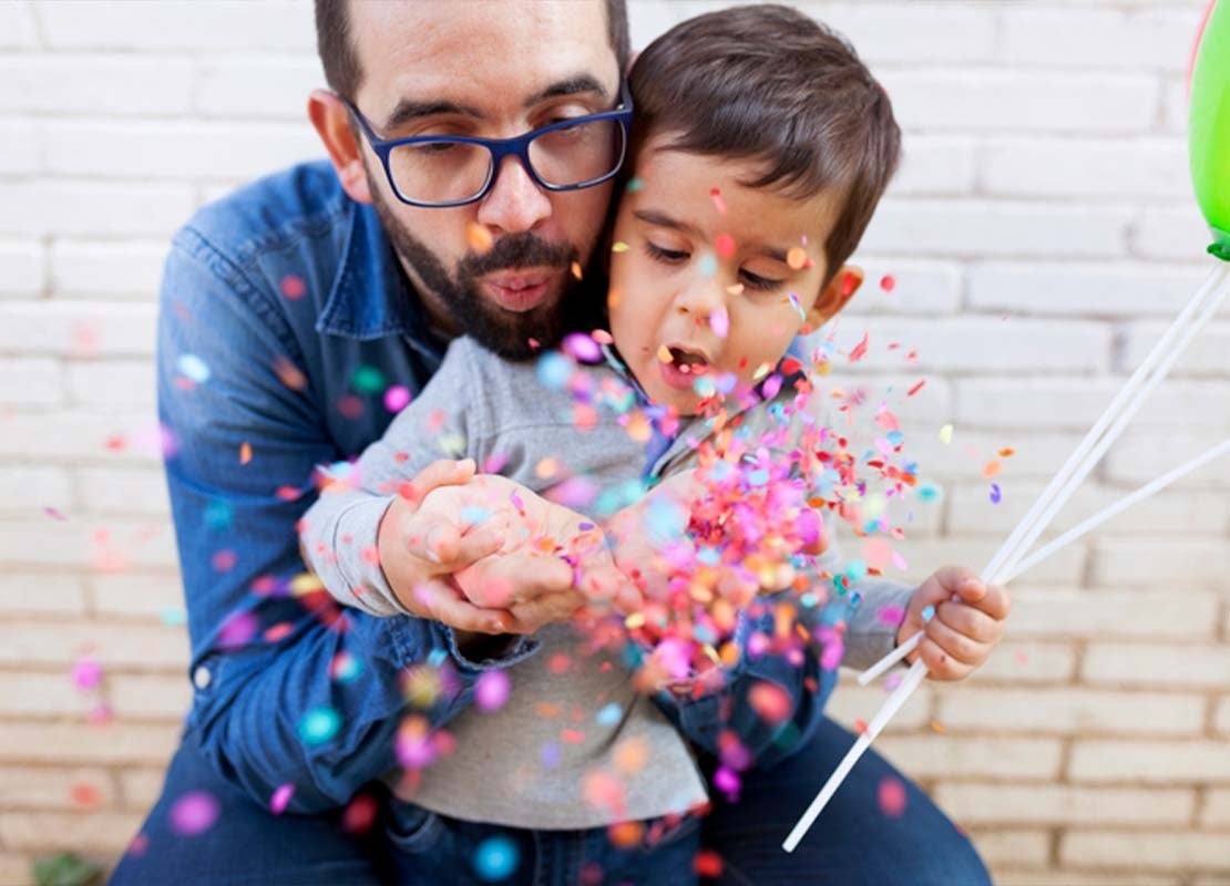 Papá sopla confeti mientras abraza a su hijo, quien sostiene un globo en el Día del Padre