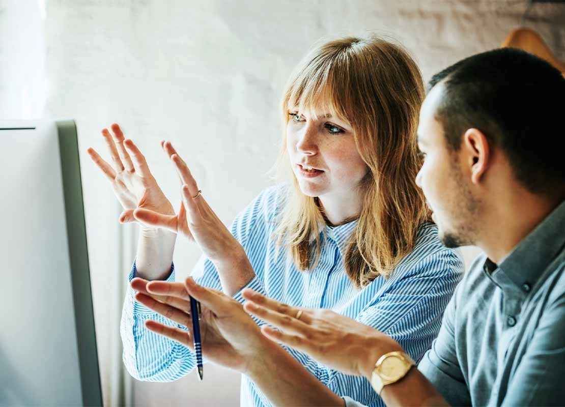 Mujer habla con su compañero mientras diseñan un estudio de mercado frente a un computador