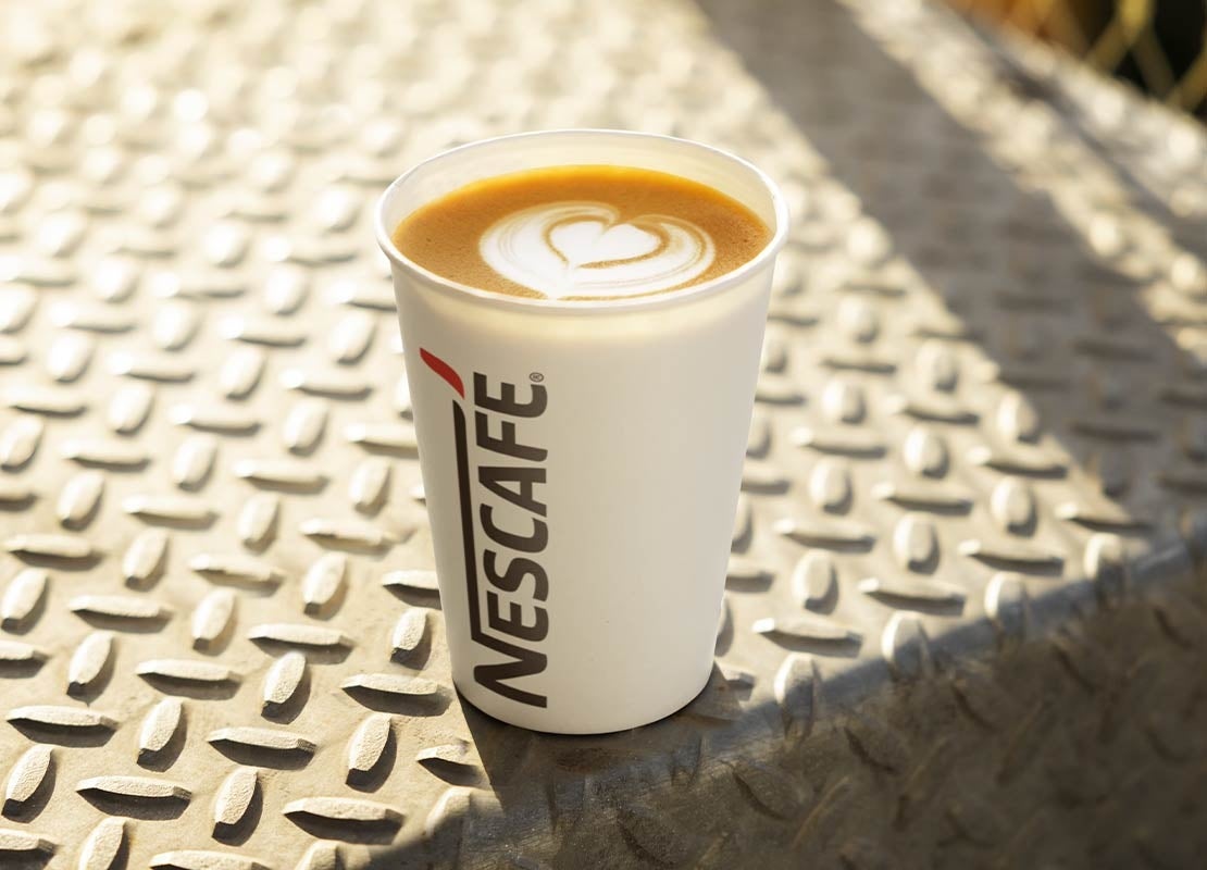 Sobre un escalón de acero, un vaso de NESCAFÉ® blanco con un café que lleva un diseño de latte art