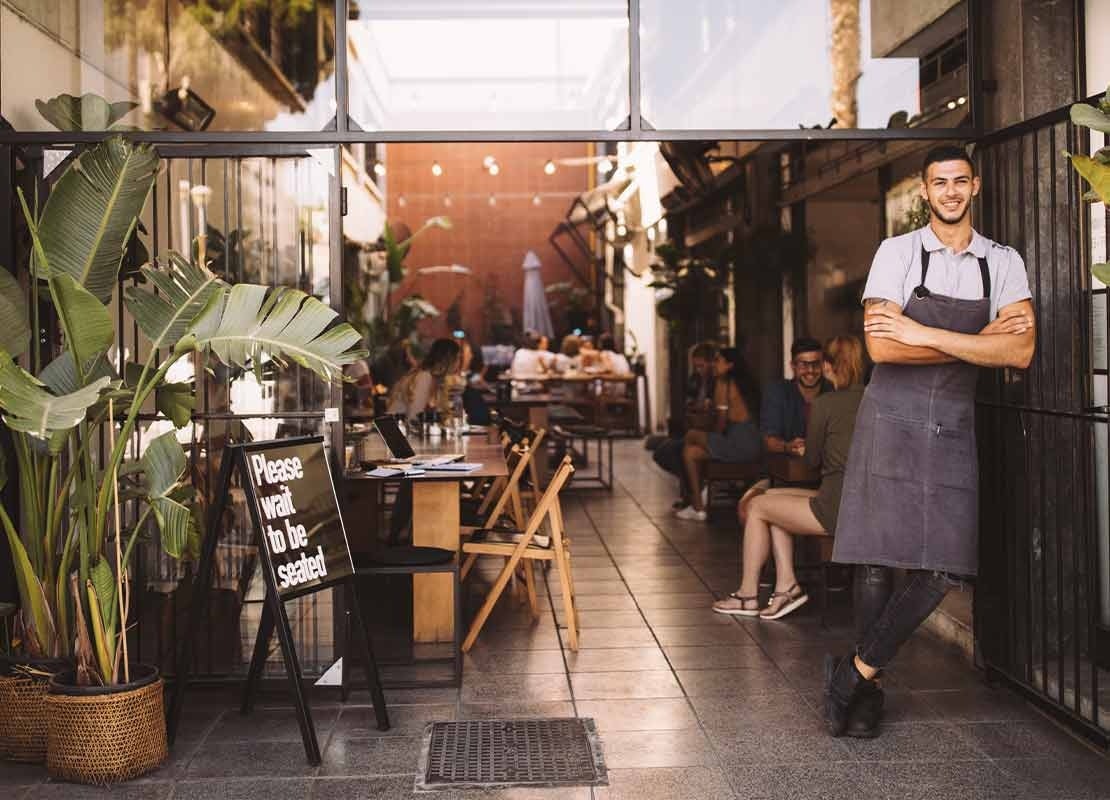 Hombre cruzado de brazos en la entrada de su negocio de comida y al fondo el mostrados y sus clientes