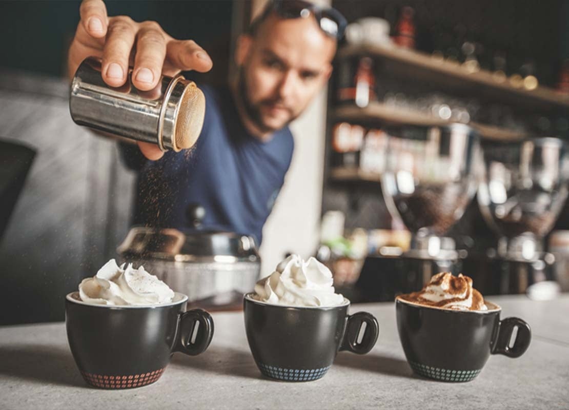 Barista espolvorea cacao sobre tres tazas de cappuccino coronadas con crema para un menú de café